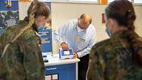 Ein Mann zeigt mit seinem Finger auf das Display eines PCR-Gerätes. Zwei Soldatinnen hören den Mann zu.