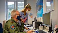 Ein Soldat sitz vor einem PC-Bildschirm und hält ein Telefon ans Ohr. Neben ihm steht eine blonde Frau mit Mundschutz.