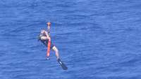 Ein Rettungsschwimmer mit Unterarmgurt an einem Windenseil, unter ihm das Meer