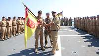 Zwei Soldaten in sandfarbener Arbeitsuniform nebeneinander und eine Soldat in Wüstentarn Uniform mittig mit rot-gelben Fahne.