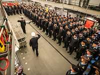 Mehrere Soldaten in blauer Arbeitsuniform stehen nebeneinader im Hangar eines Kriegsschiffs.