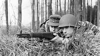 Zwei Soldaten – einer mit Mütze, einer mit Helm – liegen mit einem Gewehr im Anschlag auf dem Waldboden in Stellung.