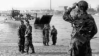 Schwarz-Weiß-Foto: Mehrere Soldaten stehen am Strand, ein Landungsboot nähert sich. 