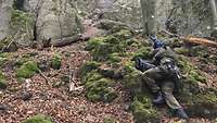 Soldaten liegen mit Gewehren im Wald vor einem Felsen.