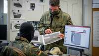 Ein Soldat sitzt am Computer, ein anderer steht vor ihm und erklärt eine ausgedruckte Grafik.
