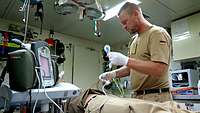 Ein Soldat steht in einem Behandlungsraum neben einer Liege mit Patient und hält mehrere Kabel eines EKG-Geräts in der Hand 