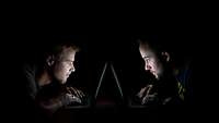 Zwei Männer sitzen sich im Dunkeln an einem Tich genüer und starren gebanntauf ihre Laptops