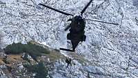 Ein Hubschrauber schwebt vor einem Berg, darunter hängen zwei Soldaten am Seil.