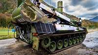 A tank with a big excavator bucket standing in front of a water basin, in the background high mountains and blue skies