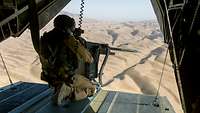 Ein Soldat an der Waffe hockt auf der Landerampe eines Hubschraubers vom Typ CH-53 während des Fluges über eine Wüstenlandschaft
