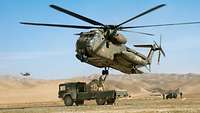 Ein Hubschrauber vom Typ CH-53 fliegt dicht über der Ladefläche eines LKWs, auf dem ein Soldat steht und etwas befestigt