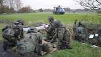 Im Vordergrund kümmern sich auf einer Wiese Soldaten um einen Verletzten. Im Hintergrund steht ein Hubschrauber auf der Wiese.