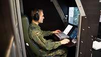 A man sits in a simulator in front of a monitor, wearing headphones, hands on a control console.