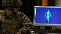 A soldier wearing a protective vest, helmet and glasses is sitting beside a blue monitor showing a simulated soldier