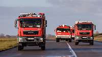 Vier Feuerwehrwagen fahren auf einer Start- und Landebahn.