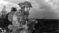 Schwarz-Weiß-Aufnahme: Ein Soldat in Flecktarn mit voller Ausrüstung, Sprechfunkgerät und Gewehr kniet im Gelände