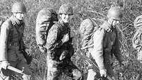 Schwarz-Weiß-Aufnahme: Soldaten in Uniform mit und ohne Flecktarn gehen hintereinander