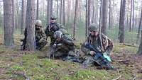 Vier Soldaten knien im Wald. Einer von ihnen gibt einen Befehl per Funk.