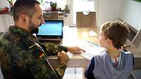 Der Vater in Uniform sitzt mit seinem Sohn am Esstisch vor einem Laptop und einem Rechenbuch.