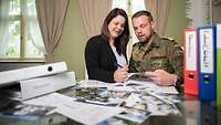 Ein Bundeswehrsoldat und eine Frau stehen an einem Tisch mit Ordner und Broschüren