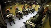Mehrere Soldaten mit Helm, Sauerstoffmaske und Notebook sitzen in einer Unterdruckkammer nebeneinander