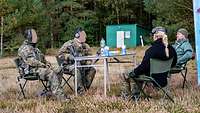 Zwei Soldaten in grüner Uniform und zwei Zivilisten sitzen draußen an einem Tisch und führen ein Gespräch.