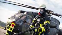 Ein Feuerwehrmann mit einer Atemschutzmaske hält einen Feuerwehrschlauch. Hinter ihm steht ein Hubschrauber.