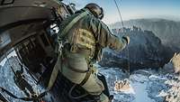 Ein Soldat steht gesichert an der geöffneten Seitentür eines Hubschraubers und hält ein Außenseil, unter ihm das Gebirge.