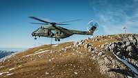 Ein Hubschrauber schwebt mit einer Außenlast über dem Boden in einem Gebirge.