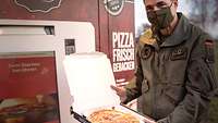 Männliche Person in Uniform präsentiert eine Pizza in einem aufgeklappten Karton