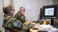 Two soldiers are sitting side-by-side at their computers. A Plexiglas screen separates their workstations