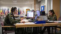 Two soldiers of MMCC/EMC are sitting face-to-face at their workstations. A Plexiglas screen separates them.