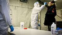 A man in protective clothing takes a nose swab from a civilian test person behind a table.