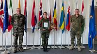 Three service men are standing in front of national flags. The female in the middle holds a coat of arms and a picture