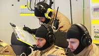 Drei Soldaten in Gefechtsanzug mit Flammschutzhaube, Flammschutzhandschuhen und Kopfhörern arbeiten konzentriert