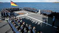 Marinesoldaten in dunkelblauer Arbeitsunifomen stehen in mehreren Reihen angetreten auf dem grauen Flugdeck eines Schiffes.