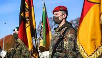 Zwei Soldaten in grüner Uniform stehen neben der deutschen Flagge auf einem Appellplatz.