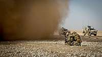 Zwei Soldaten hocken am Boden, vor ihnen eine riesige Staubwolke. Im Hintergrund stehen zwei gepanzerte Fahrzeuge.