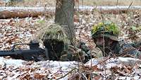 Zwei Soldaten liegen im leicht verschneiten Wald, der linke mit Gewehr im Anschlag, der rechte weist ihn mit Fernglas ein.