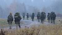 Bei nebligem Wetter marschiert eine Gruppe Soldaten mit Gepäck, Waffen und Ausrüstung auf einer unbefestigten Straße.