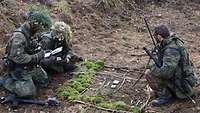Drei bewaffnete Soldaten hocken im Wald um eine aus Stöcken, Moos und Kleinteilen improvisierte Lagekarte.