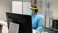 Eine Ärztin im Karrierecenter mit Gesichtsvisir, Mund-Nasenschutz und Schutzkleidung sitzt an ihrem Arbeitsplatz