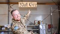 Ein Soldat sitzt in einer Lagerhalle und zeigt auf ein Holzschild mit der Aufschrift „Material Gruppe“