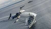 Ein kleines Jetflugzeug fliegt über ein graues Kriegsschiff in See.