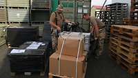 Zwei Soldaten stehen zwischen Metallkisten und Kartons und packen das eingetroffene Material aus