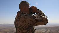 Ein Soldat im Profil blickt durch ein Fernglas auf eine Wüstenlandschaft
