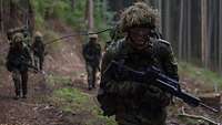 Ein Soldat mit Helm, Waffe und Marschgepäck marschiert mit seinen Kameraden durch den Wald.