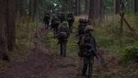 Eine Gruppe von Soldaten marschiert mit Gepäck eine Anhöhe im Wald hinauf, ein Soldat dreht sich um.