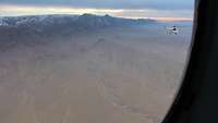 Aus dem Seitenfenster eines Hubschraubers sieht man Wüste, Gebirge und einen weißen Hubschrauber