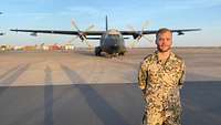 Ein Soldat steht auf dem Flugfeld und lächelt in die Kamera, im Hintergrund steht ein Transportflugzeug